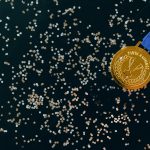 Close-up of a gold medal with blue ribbon and confetti on a black surface, symbolizing victory.